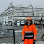 A woman wearing bright orange protective clothing standing on a construction site in snowy weather