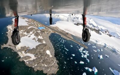 Aerial view of a polar research station with ice capped mountains and sea below and a bright blue building and a runway below