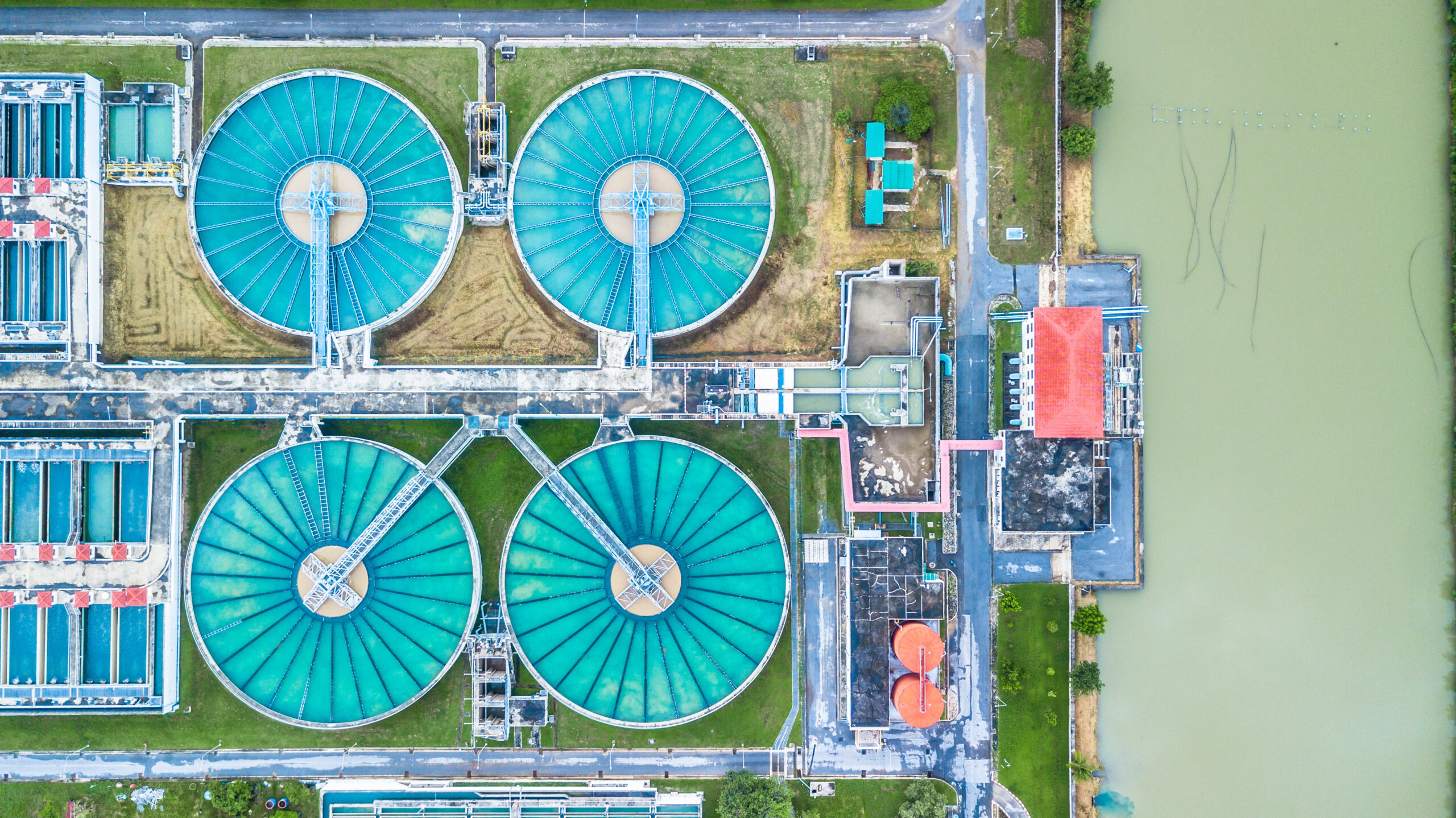 Aerial view of a water treatment plant with four large circular tanks, adjacent buildings, and a nearby body of water.