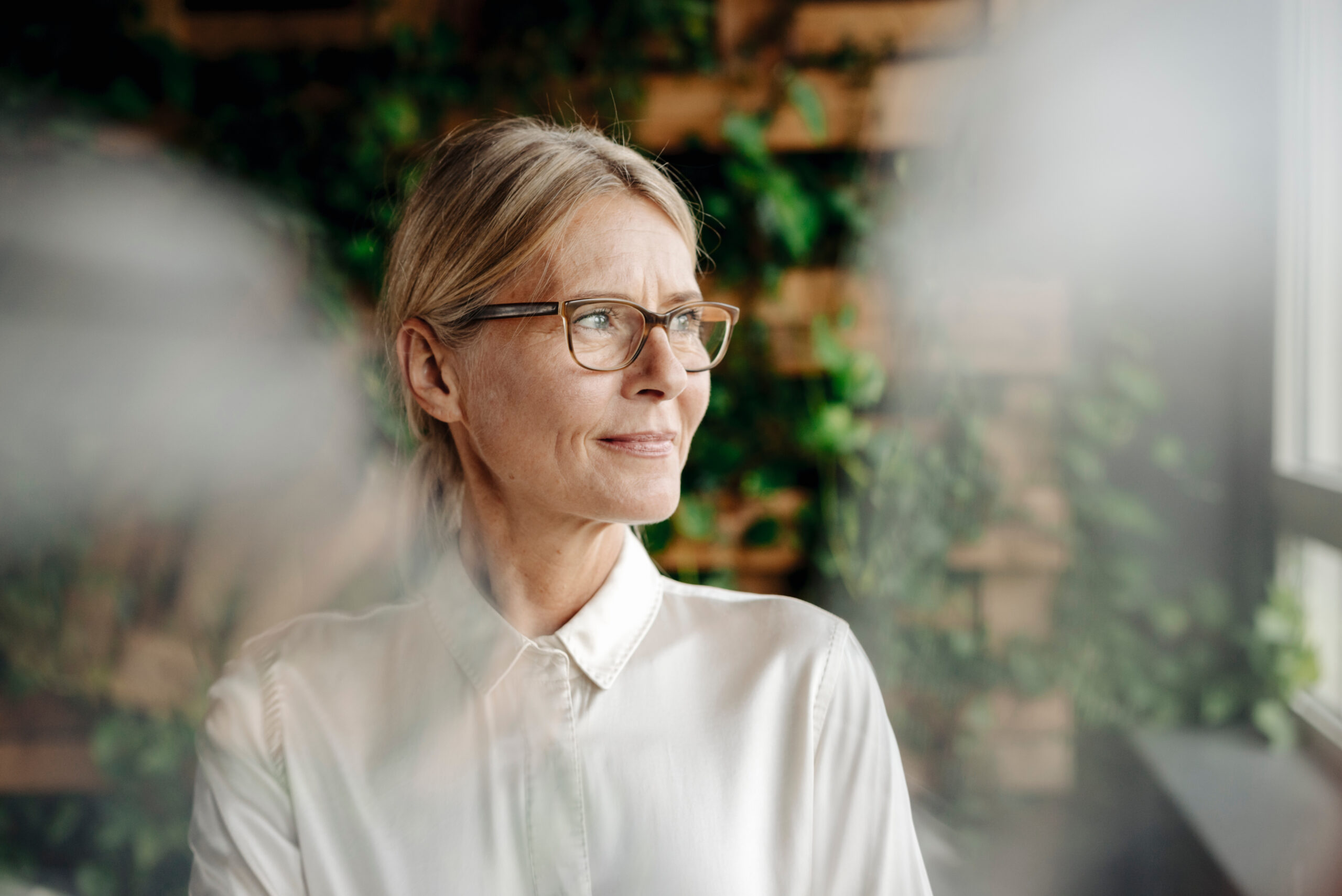 Eine ältere Frau mit Brille und weißer Bluse blickt aus dem Fenster, vor einem Hintergrund aus grünem Laub und Backsteinen.