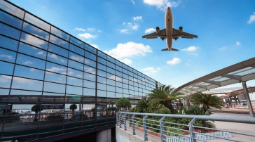 Airplane flying over a modern airport terminal with glass exterior, blue sky, and palm trees.