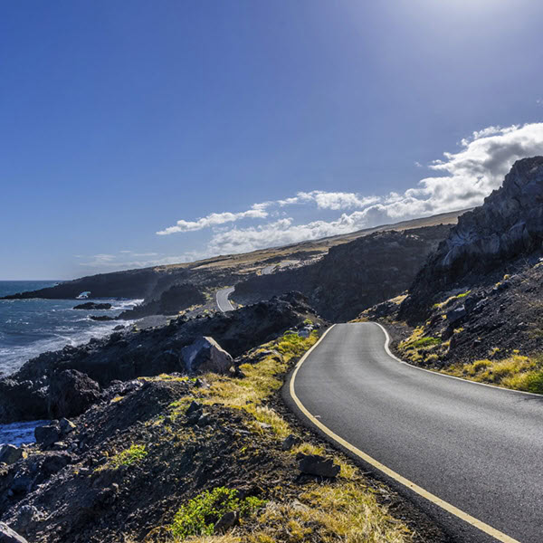 A winding road along a rocky coastal landscape with the ocean on the left and a clear blue sky above, partially covered by white clouds, could easily be a view managed by the Hawaii Department of Transportation.