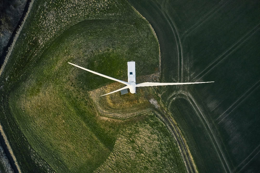 An aerial view captures the impressive infrastructure of a wind turbine situated in the middle of a green field, its towering presence casting a shadow over potential transportation routes.