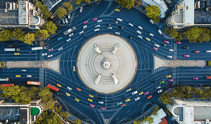 Aerial view of a circular intersection with heavy traffic around a central monument, surrounded by trees and buildings.