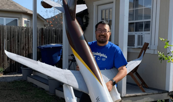 Un homme se tient dehors, tenant une grande maquette d'avion à côté d'une petite maison avec une terrasse en bois.