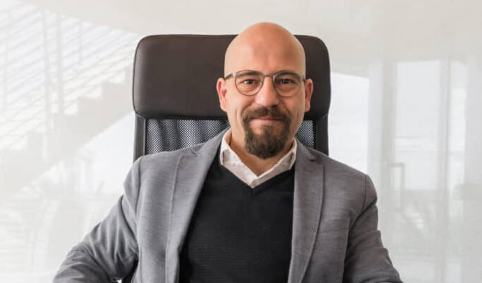 Un homme avec des lunettes, une barbe et une moustache est assis sur une chaise de bureau, portant un blazer gris, un pull noir et une chemise blanche.