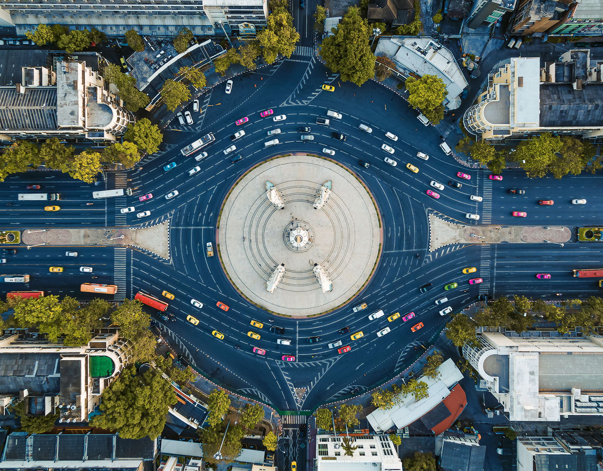 Eine Draufsicht auf einen belebten städtischen Kreisverkehr mit einem großen kreisförmigen Denkmal in der Mitte. Der Kreisverkehr ist von mehreren Fahrspuren umgeben, auf denen sich bunte Autos, Busse und Taxis bewegen. Bäume und Gebäude bilden den Rahmen der Szene und unterstreichen die lebendige städtische Umgebung.