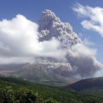 A dark coud of ash rises over the Sofrier Hill volvano in Montserrat. The volcano is obscured by white clouds and there are green hills in the foregrund.