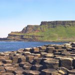 The Giant's Causeway in Northern Ireland. Hexagonal blocks of brownish grey rock in the foregroud, with seain the biddle ground and a high cliffs of basailt columns and green grass in the background.