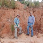 Two white men are standing in front of a low, sandy cliff