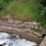 A low cliff with reddish and white layers of rock beside foaming river water. A person wearing a green coat is examining the rocks. There are trees and grass on the slope above the cliff.