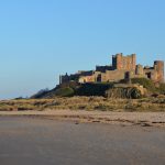 A castle sits on top of a high outcrop of rock. There and sand dunes and a beach in front of it.