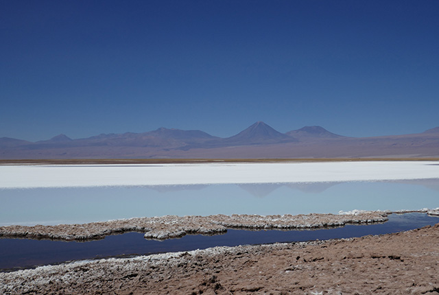 A dry, hot, flat desert with a lake and white salt flats in the foreground and mountains in the background.