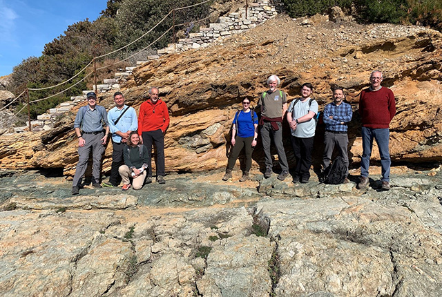 Nine people in two groups (four to the left; five to the right) standing on grey rocks with brown cliffs behind them.