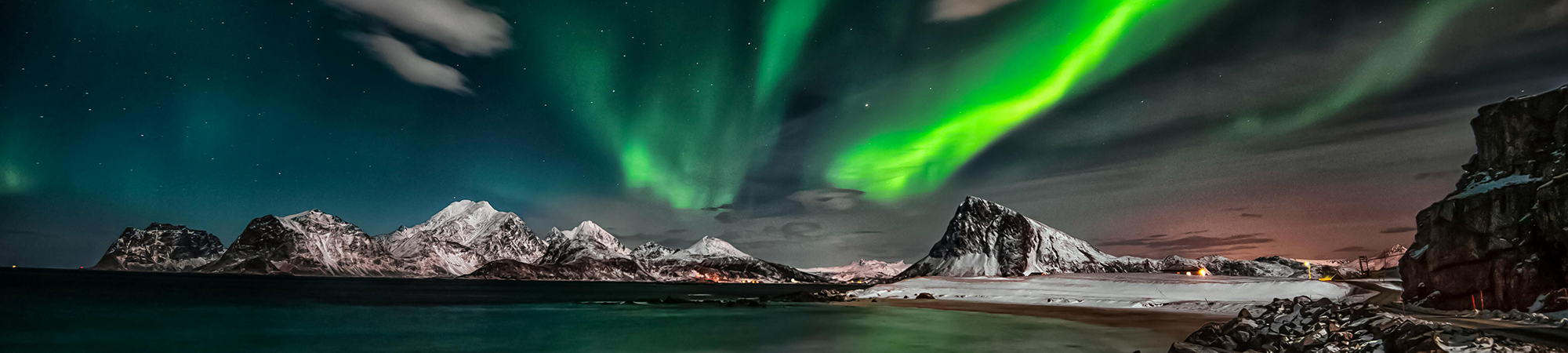 Green ribbons of aurora in the dark sky over snow covered mountains, which surround a bay