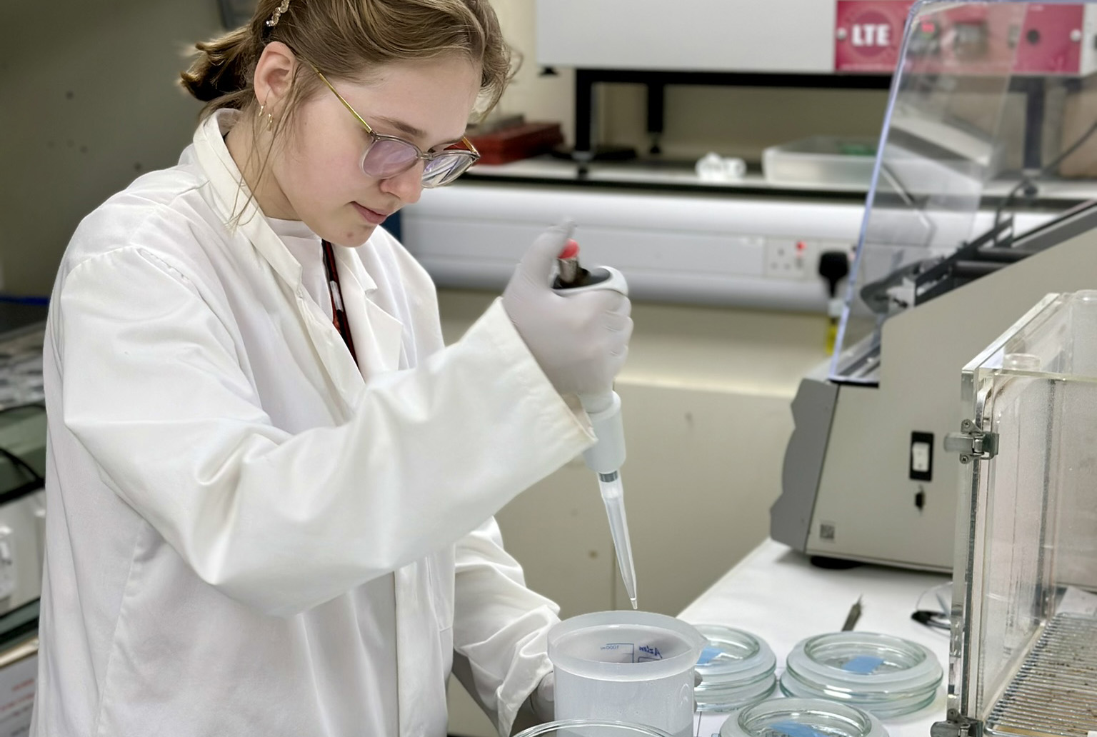 A woman in a lab coat, gloves and goggles uses a pipette to add some liquid to a container