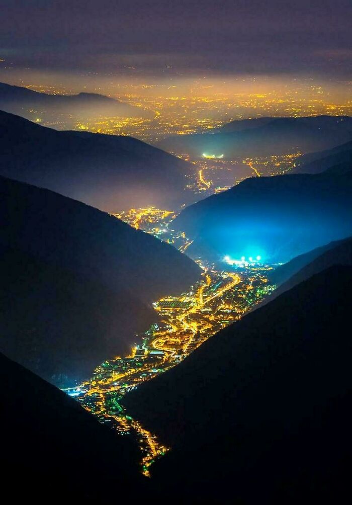 Nighttime aerial view of a vast cityscape nestled between mountains, illuminated by city lights, showcasing megastructures.