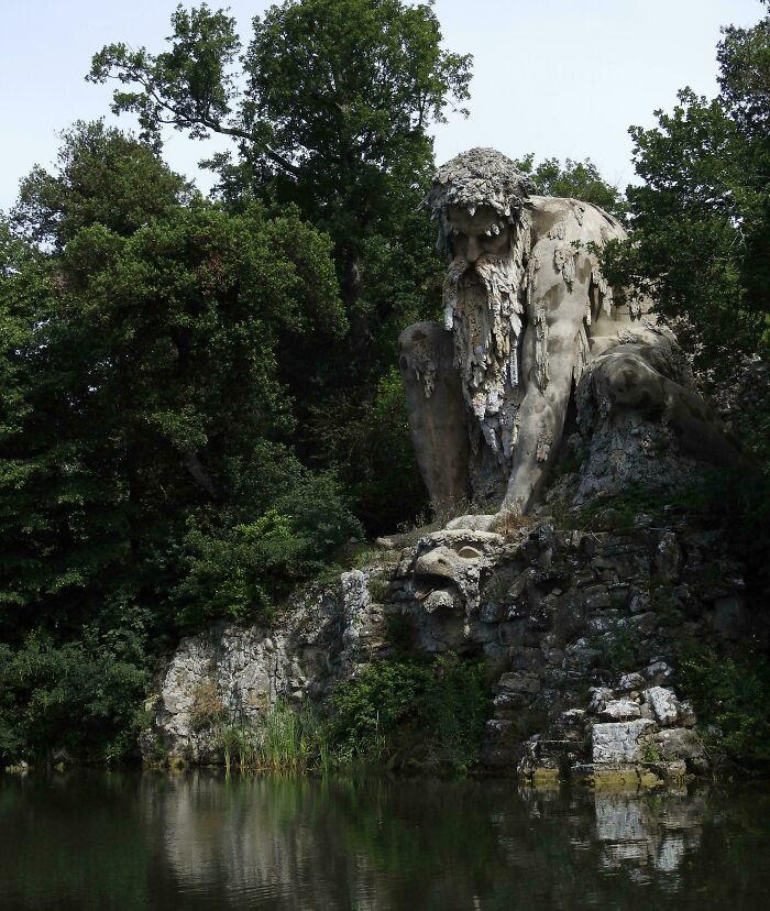 Gigantic stone statue resembling a giant, surrounded by lush greenery, reflecting on a calm water surface.