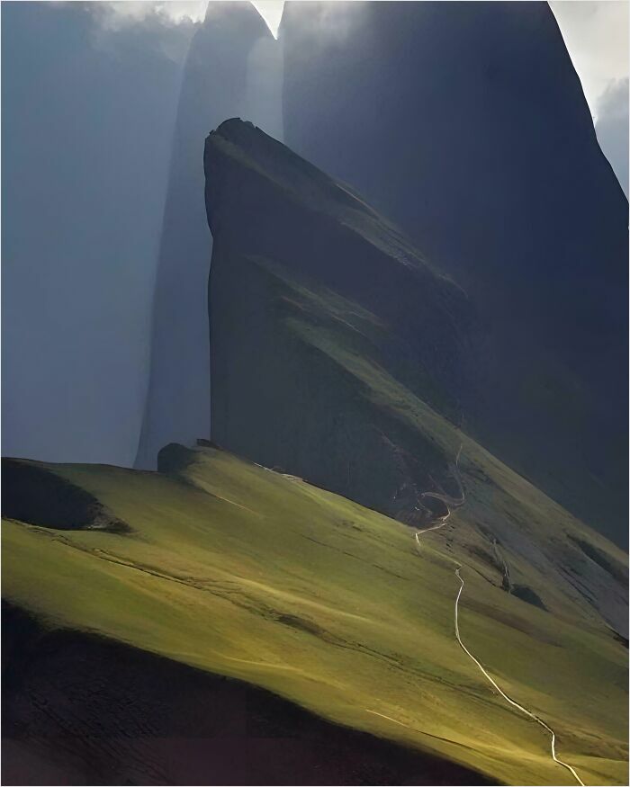 Gigantic mountain with steep, towering peaks casting shadows on the grassy landscape below, invoking a sense of megalophobia.