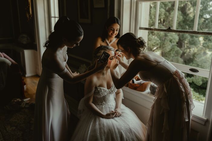 Bridesmaids help a bride get ready, capturing a top 2024 wedding moment by a window.