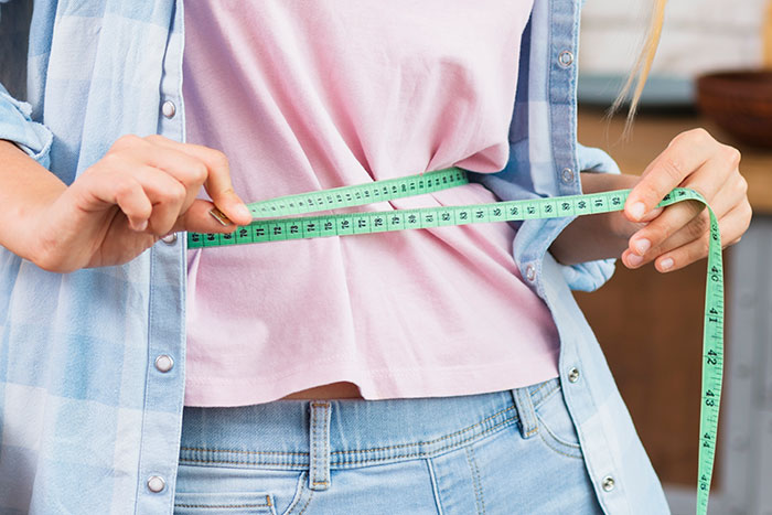 A woman measures her waist with a green tape, wearing a pink shirt and denim jeans.