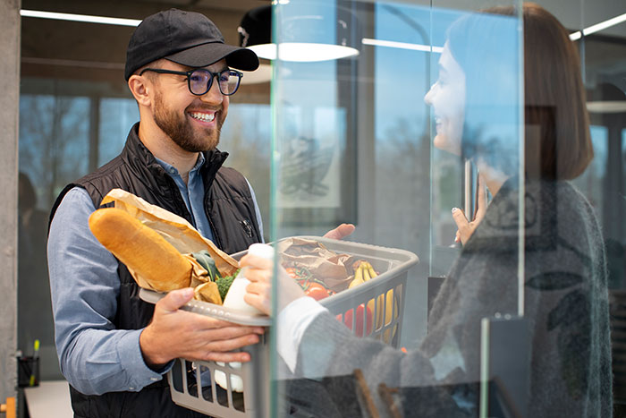 Man smiling and handing groceries to a woman through a glass door, capturing a moment of kindness and good karma.