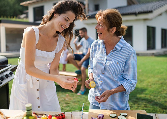 Two women smiling while preparing food outdoors; a relaxed gathering vibe.