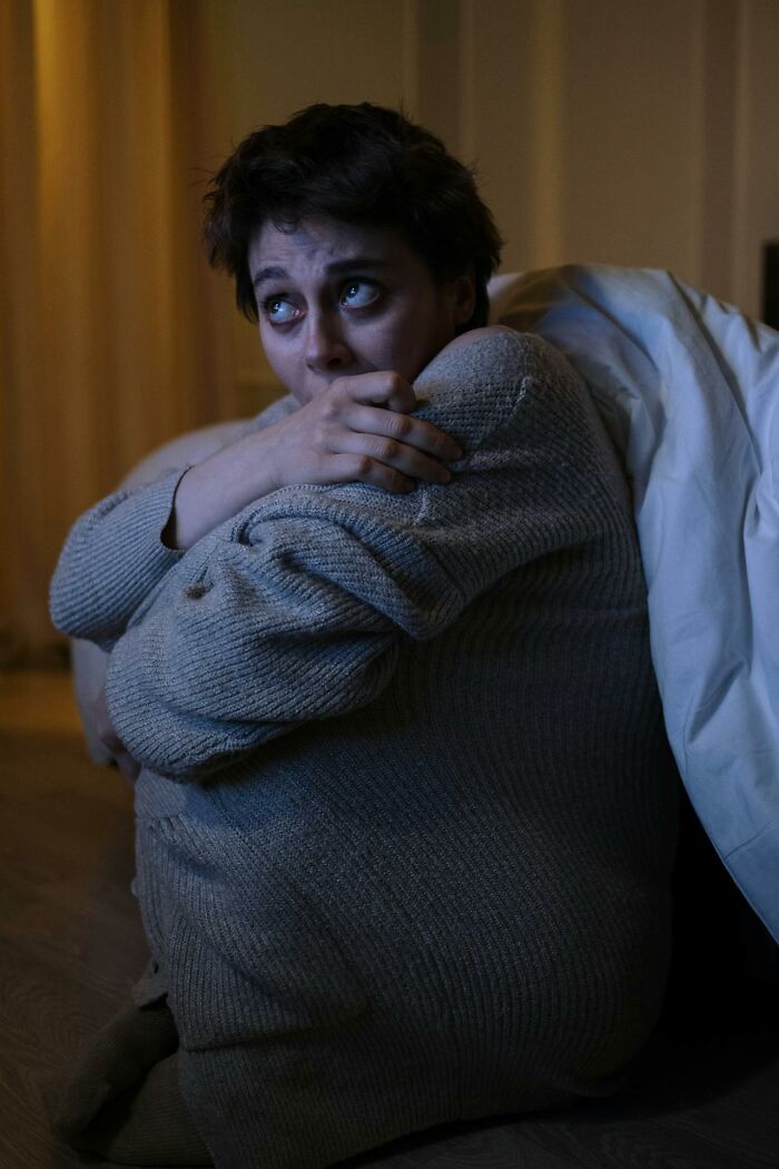 Person in a dimly lit room, wrapped in a sweater, looking anxious, reflecting behind-the-scenes casino employee experiences.