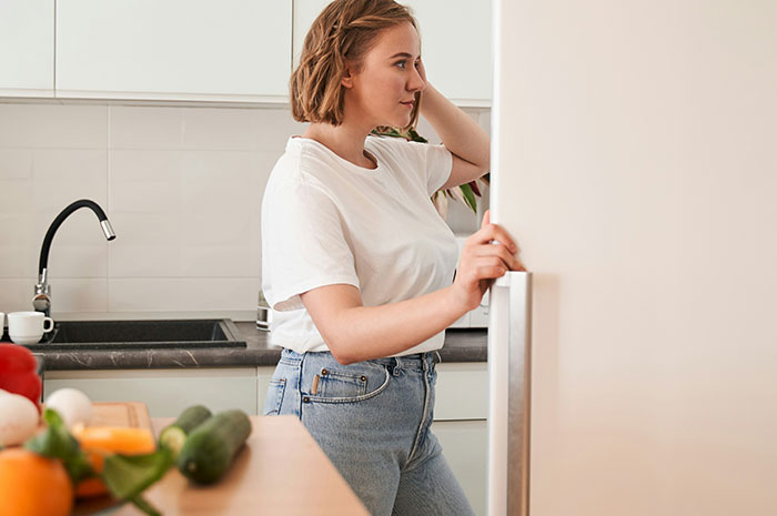 Roommate checks fridge, unaware of surprise trap set in kitchen with vegetables on counter.