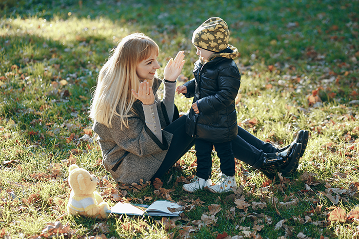 Woman sitting on grass playing with a toddler, surrounded by autumn leaves.