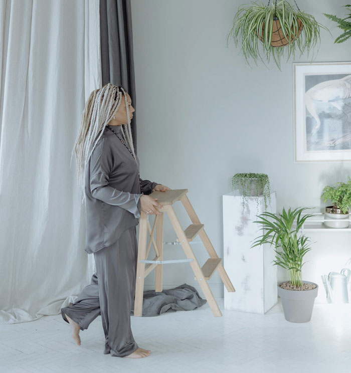 Woman in gray outfit holding a step ladder in a modern, plant-filled living room.