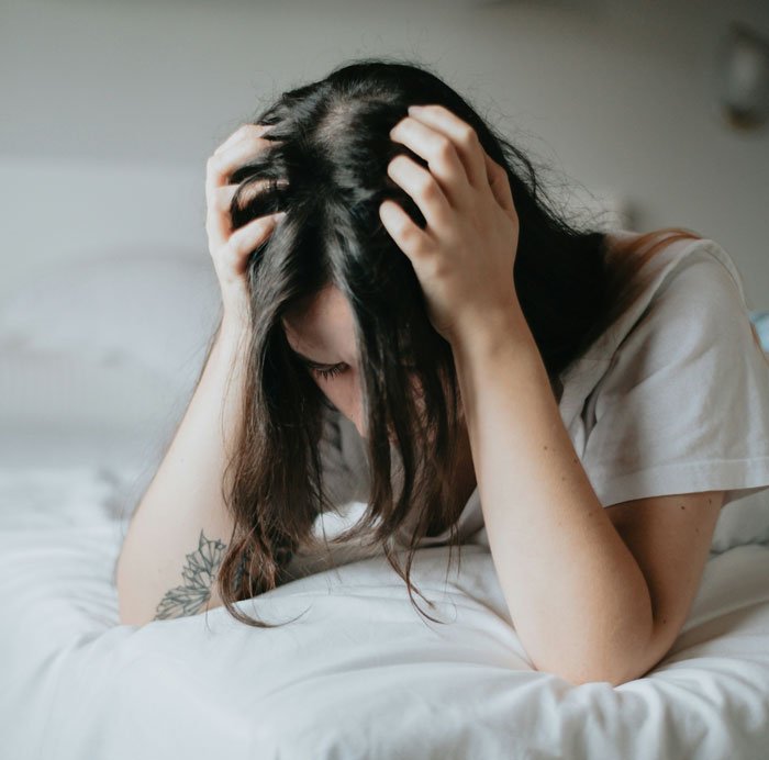 Woman stressed, holding her head, on a bed, pondering financial strain from not buying a friend's replacement hairdryer.