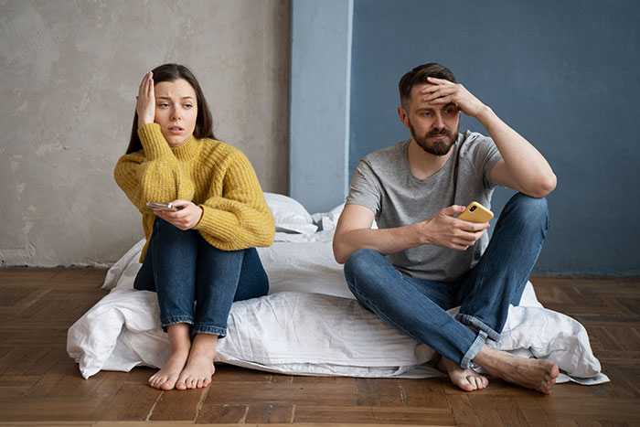 Couple sitting on bed, both looking distressed, discovering hidden personality traits, holding smartphones.