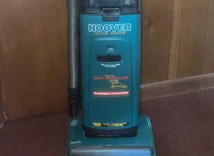 Vintage Hoover vacuum cleaner being used the wrong way, placed upright against a wall on a carpeted floor.