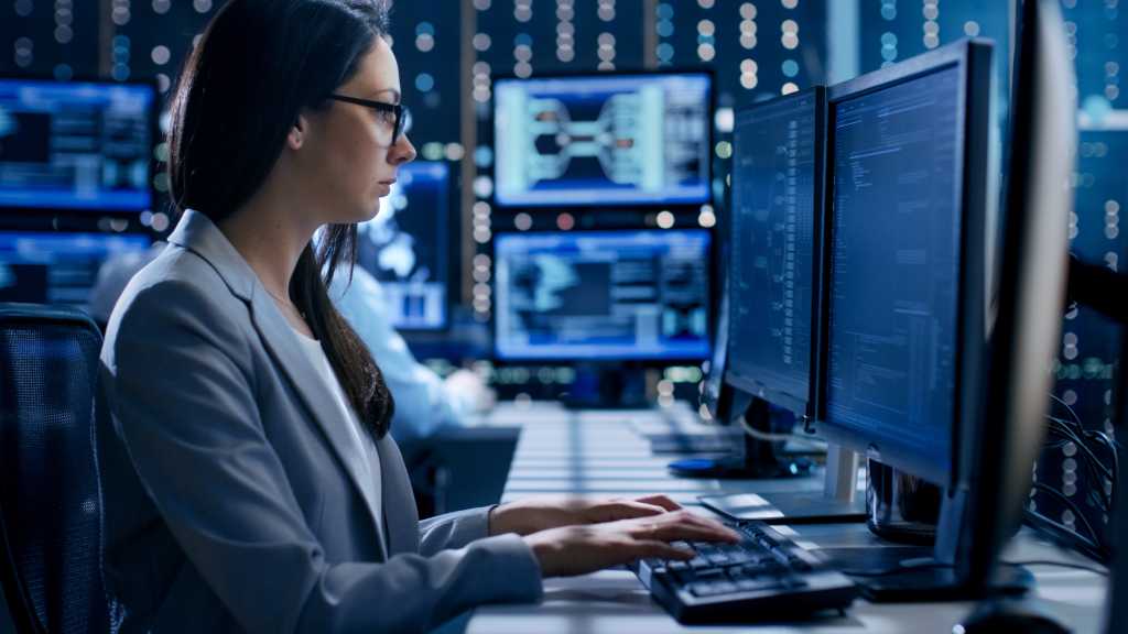 Female Engineer Controller Observes Working of the System. In the Background People Working and Monitors Show Various Information.