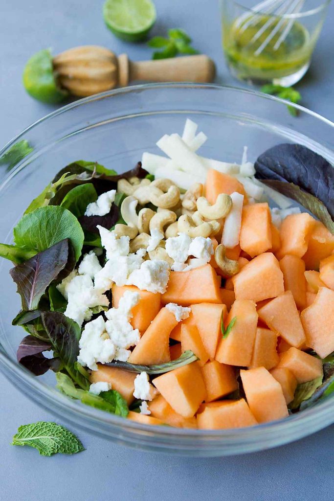 Cantaloupe, goat cheese, lettuce and cashews in a glass bowl.