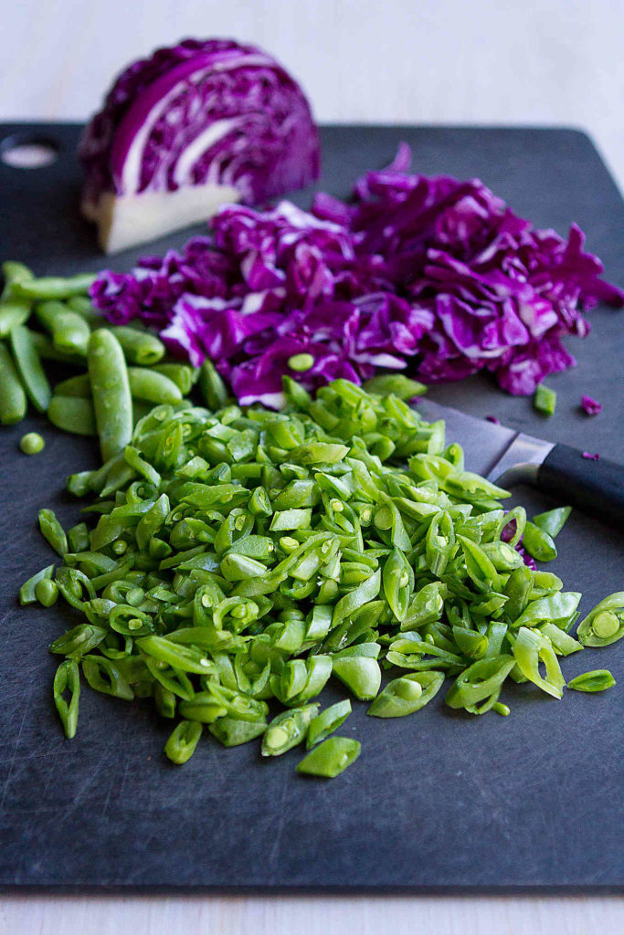 Thinly sliced snap peas and purple cabbage on a cutting board.