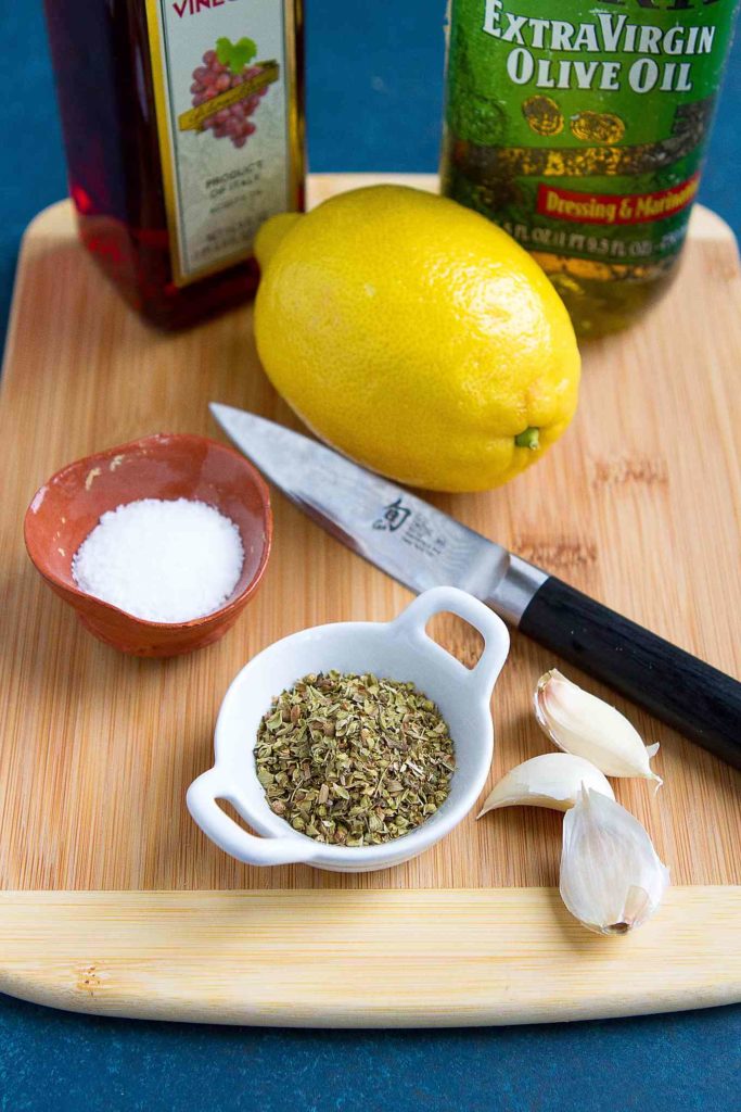 Lemon, oregano, olive oil, red wine vinegar, garlic and salt and a light wood cutting board.