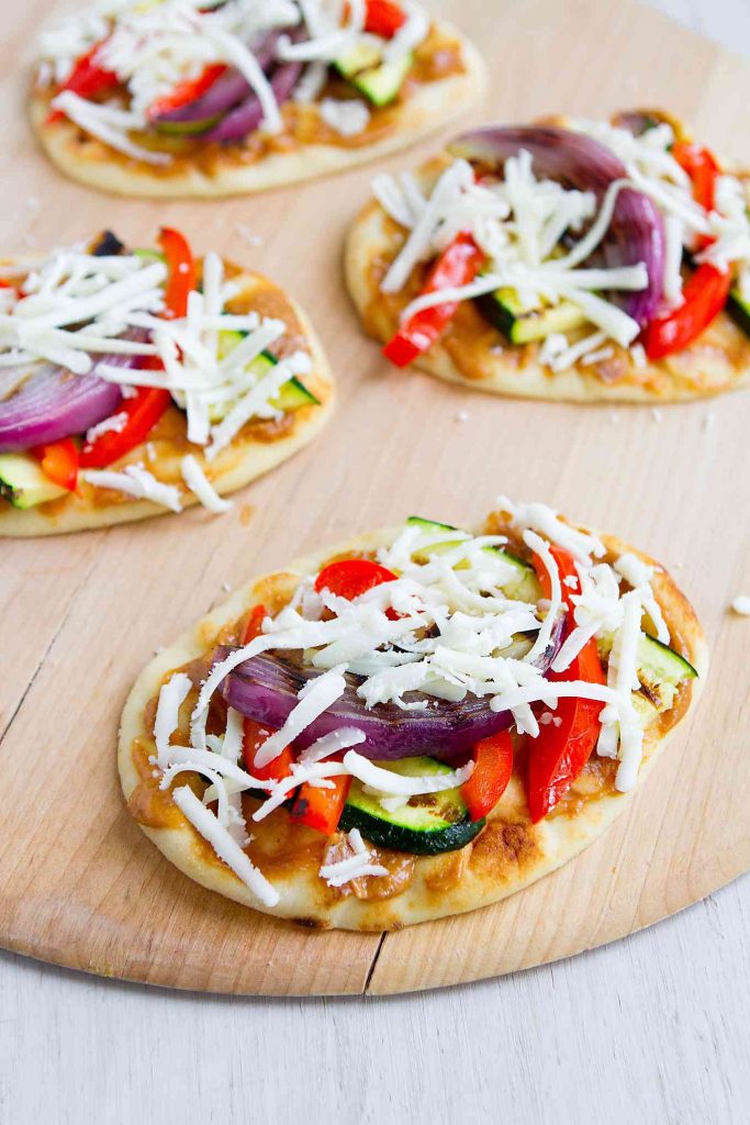 Naan bread on a pizza peel, topped with grilled vegetables, peanut sauce and a grated mozzarella cheese.