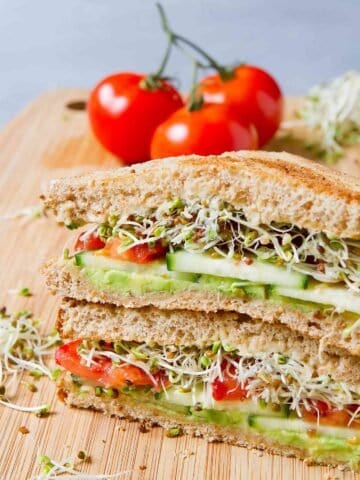 Two stacked sandwich halves, filled with avocado, cucumber, tomato and sprouts. Tomatoes in the background.