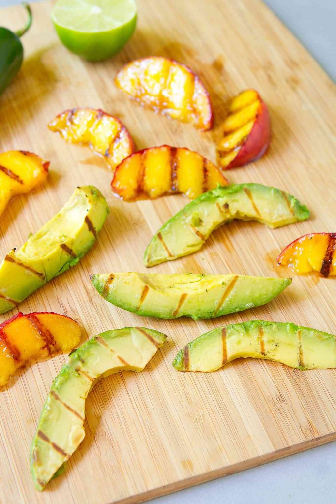 Slices of grilled avocado and grilled peach on a bamboo cutting board.