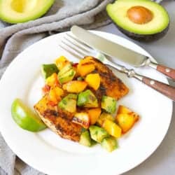 Grilled fish with avocado peach salsa on a white plate, plus two half avocados in background.