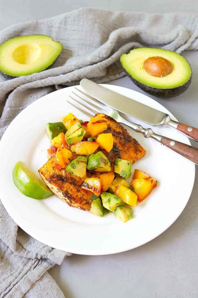 Grilled fish with avocado peach salsa on a white plate, plus two half avocados in background.