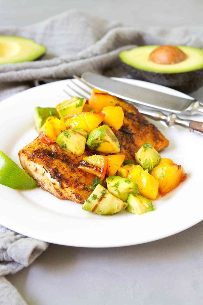 Grilled mahi mahi with avocado peach salsa on a white plate, plus two half avocados in background.