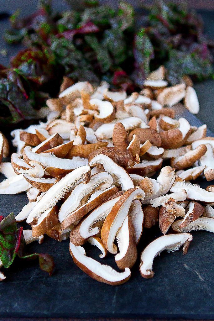 Sliced shiitake mushrooms and chopped red chard on a black cutting board.