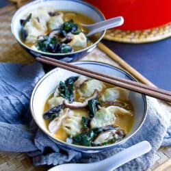 Wonton soup with mushrooms and chard in bowls. Spoons and chopsticks on the side.