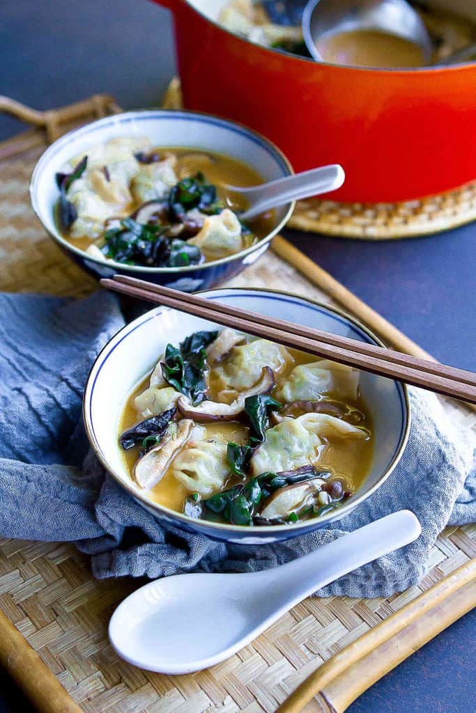 Wonton soup with mushrooms and chard in bowls. Spoons and chopsticks on the side.