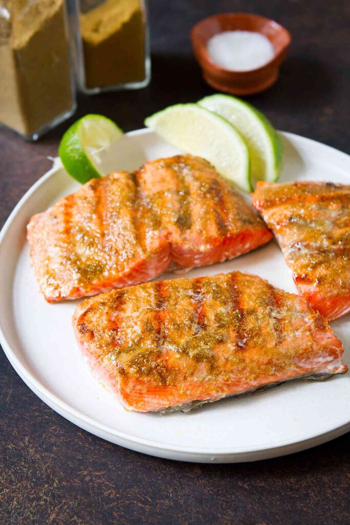 Three grilled salmon fillets on a white plate with lime wedges.