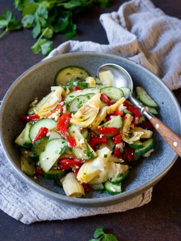 Cucumber, roasted red peppers and artichokes in a gray bowl, with parsley in background.