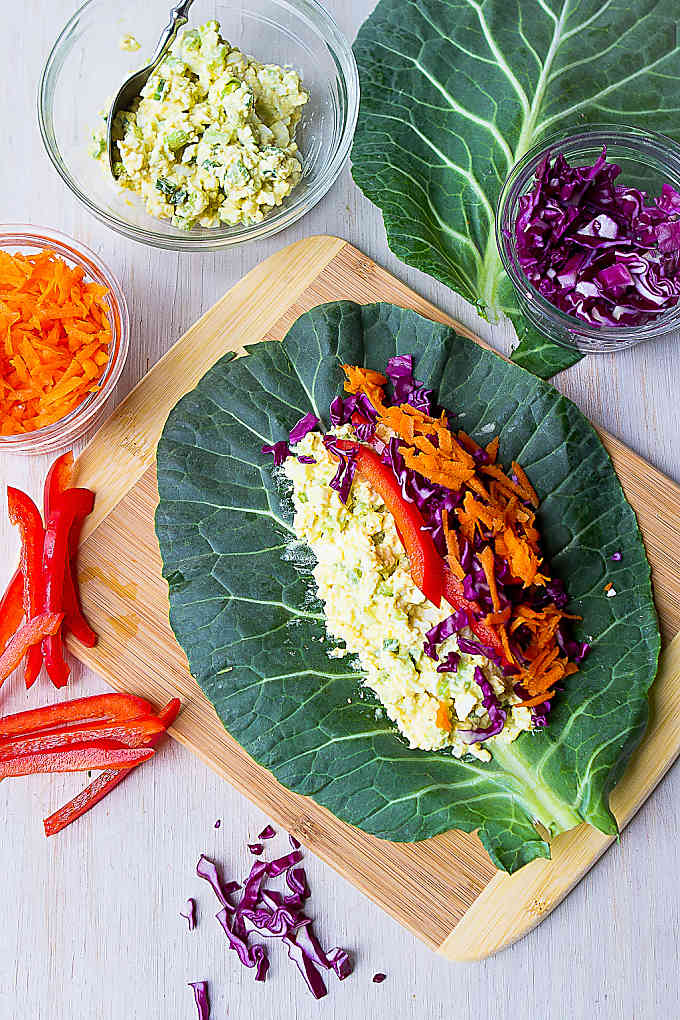 Collard green leaf topped with egg salad and vegetables, on a bamboo cutting board.